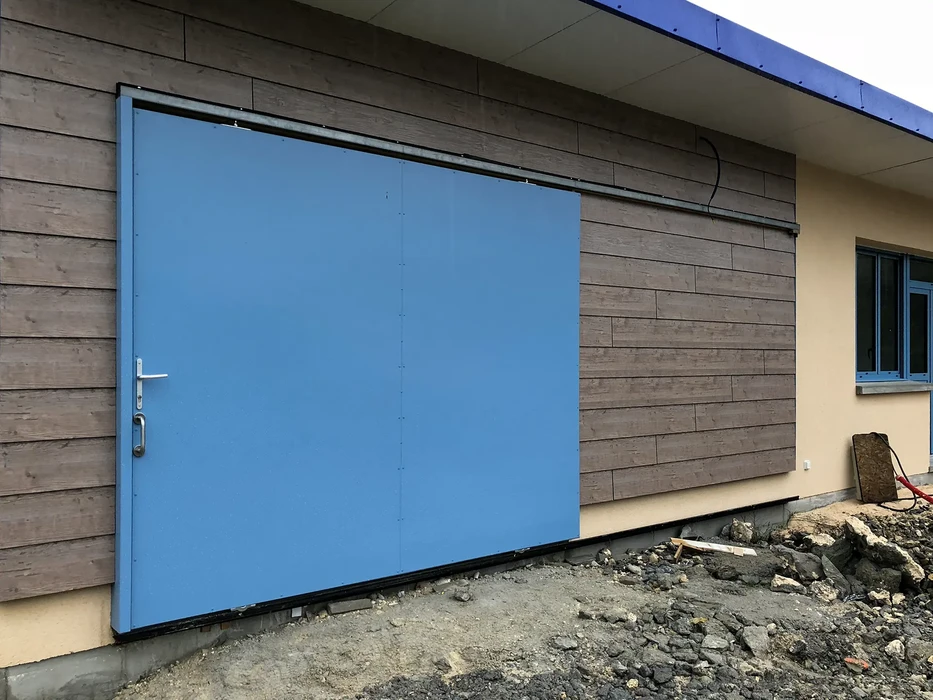 Extérieur d'un bâtiment en construction avec grande porte coulissante bleue installée sur bardage bois horizontal