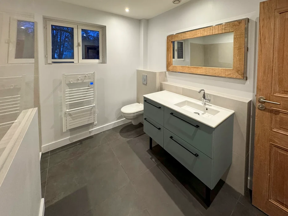 Salle de bain moderne en cours de finition avec baignoire d'angle blanche, meuble vasque suspendu en bois clair, miroir avec éclairage LED intégré