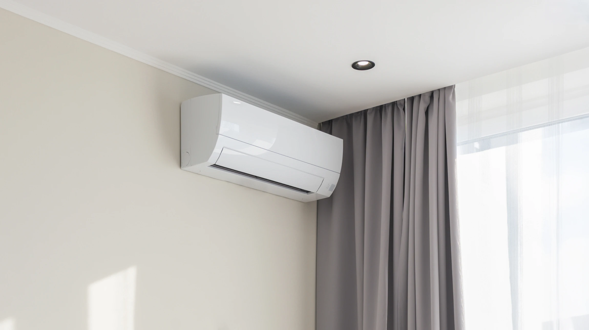 Main tenant une télécommande blanche pointée vers un climatiseur mural dans une pièce moderne avec fenêtres et rideaux clairs